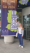 A woman is standing in front of a conference entrance next to an advertising banner.