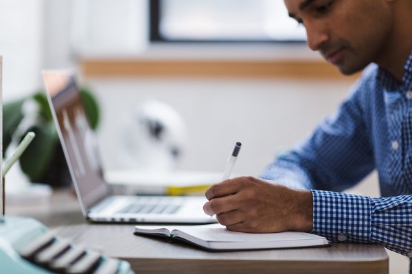 A man is sitting at a desk writing something down, with a laptop in front of him.