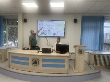 Two men on a podium. One man points to a presentation running on a screen. Another sits in front of a computer screen. 
